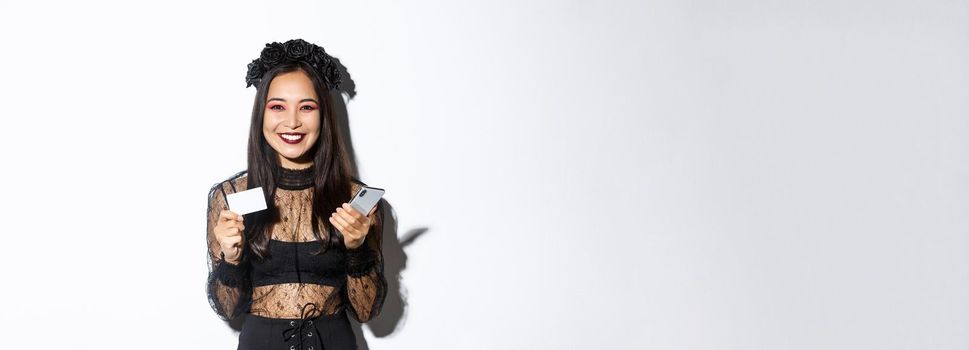 Beautiful stylish asian woman in halloween costume holding mobile phone with credit card and smiling, standing over white background.