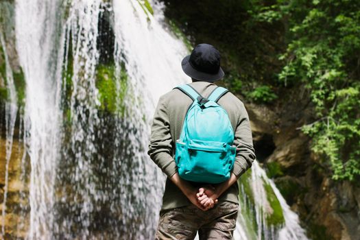 Guy in a black panama hat and with a blue backpack on his back looks at a beautiful waterfall. Foreshortening from the back. Portrait without a face.