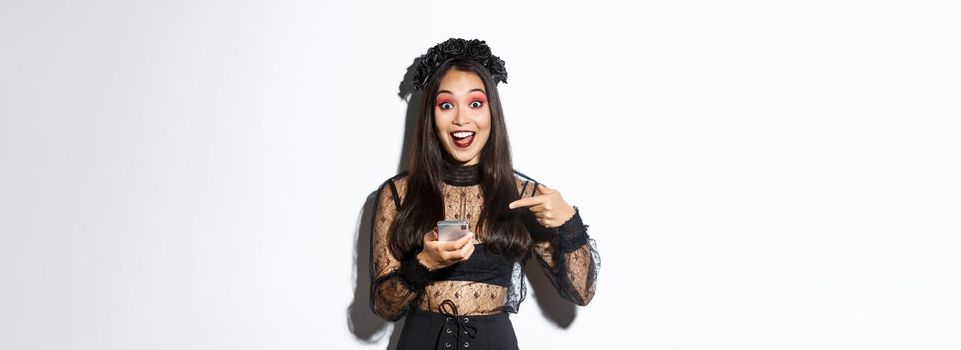 Portrait of excited asian girl pointing finger at smartphone, showing something about halloween in internet, standing over white background in gothic lace dress.