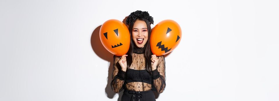 Image of asian girl in evil witch costume holding two orange balloons with scary faces, celebrating halloween, standing over white background.