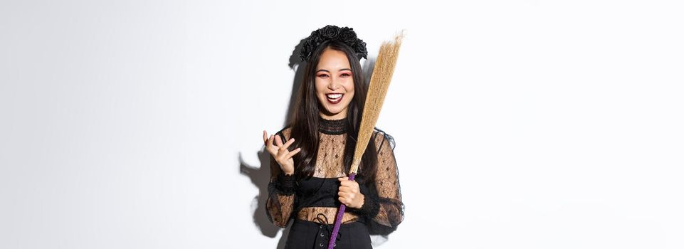 Image of witch in gothic lace dress and black wreath making evil laugh and holding broom, celebrating halloween, standing over white background.