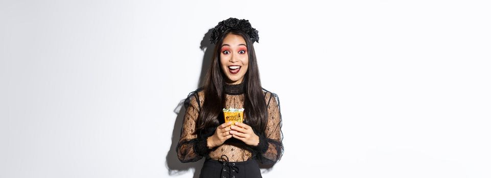 Beautiful asian girl smiling happy, holding sweets, wearing witch costume on halloween, enjoying trick or treating.
