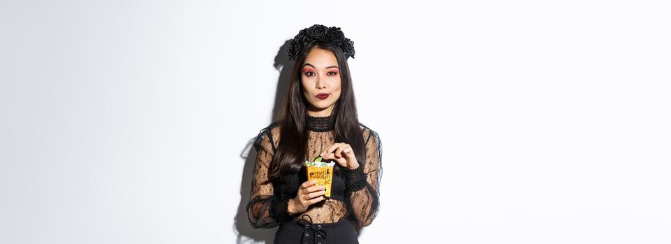 Confident smiling asian woman looking satisfied, holding sweets and wearing witch costume on halloween, standing over white background.