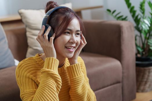 A portrait of a young Asian woman with a smiling face wearing a pair of headphones and using computer and listening to music while sitting on the sofa.
