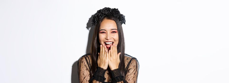 Close-up of happy asian woman celebrating halloween in witch costume and laughing, standing over white background.
