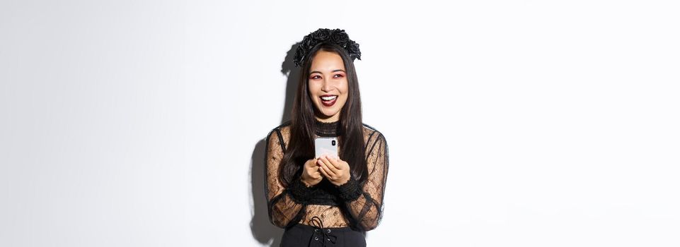 Happy attractive asian woman in halloween costume laughing carefree, holding smartphone and looking amused at upper left corner, standing over white background.