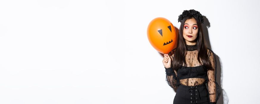 Image of silly beautiful asian woman in witch costume, holding orange balloon with scary face, celebrating halloween.