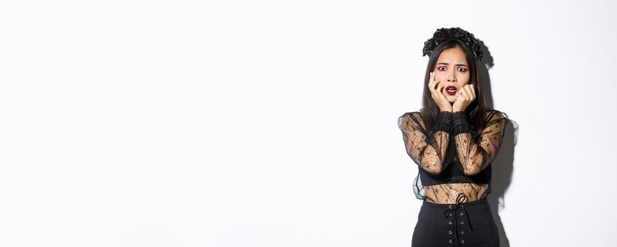 Image of scared and shocked asian female in halloween costume looking frightened at something scary, standing over white background.