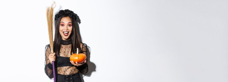 Excited attractive asian girl celebrating halloween, wearing witch costume, holding pumpkin and broom, going trick or treat, standing over white background.