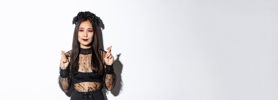 Image of hopeful asian girl in gothic lace dress and wreath making wish, cross fingers for good luck, looking wishful at camera, standing over white background.