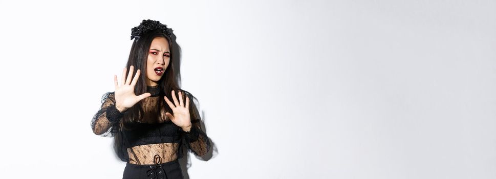 Image of bothered and annoyed asian woman in elegant gothic dress raising hands defensive, grimacing from camera flesh, asking to stop taking pictures of her, standing white background.