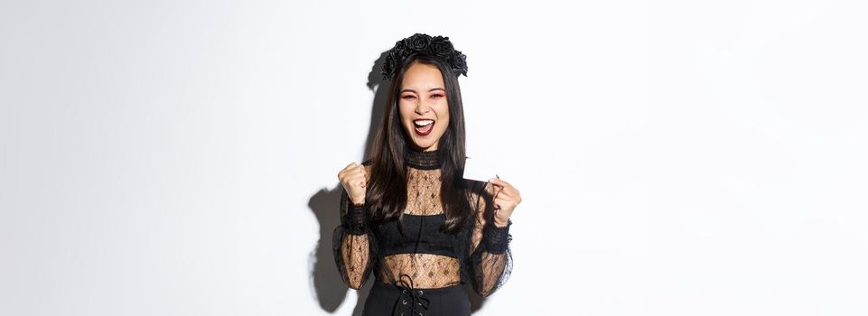 Cheerful asian woman in gothic lace dress and black wreath showing credit card and rejoicing, standing over white background.