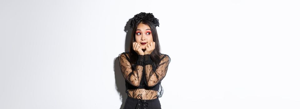 Silly beautiful asian girl in gothic lace dress with wreath looking dreamy left, have nostalgic thoughts, standing in halloween costume over white background.