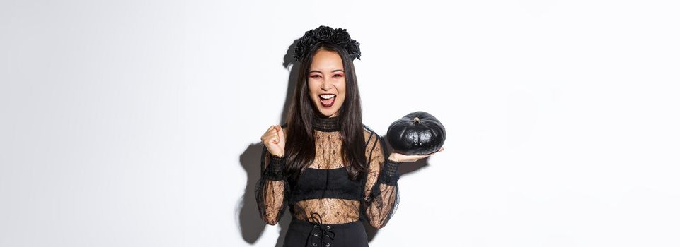 Attractive joyful asian girl enjoying halloween, holding black pumpkin and cheering, wearing witch costume, standing over white background.