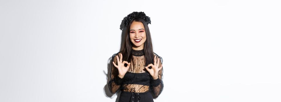 Cheerful beautiful asian woman in witch dress showing okay gestures and smiling satisfied, approve halloween costume or advertisement, standing over white background.