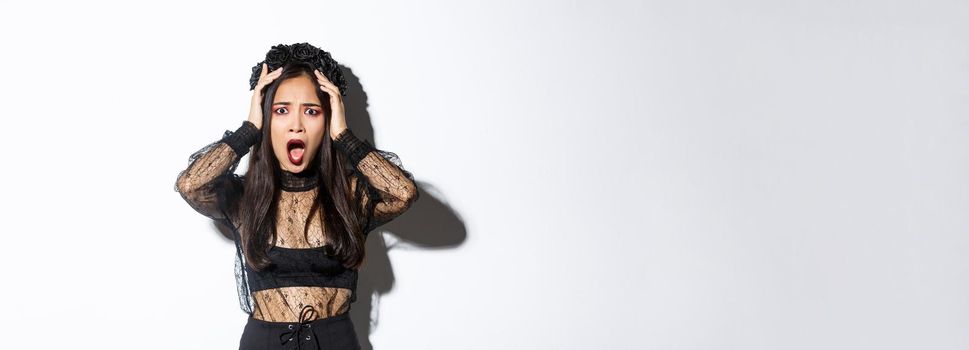 Image of horrified and shocked asian woman in gothic lace dress and wreath looking ambushed, wearing halloween costume, gasping and looking concerned, standing over white background.