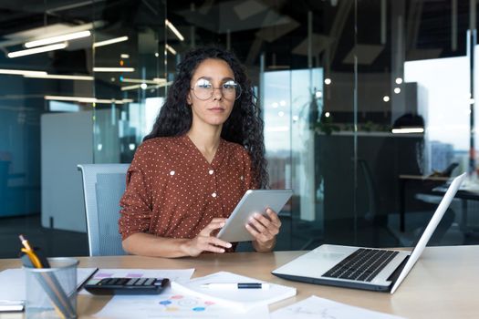 Thinking businesswoman with tablet computer looking seriously at camera, Hispanic woman working inside modern office building with documents