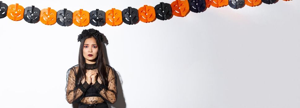 Image of scared and worried asian woman in witch costume looking concerned, wearing witch costume and standing against pumpkin banners.