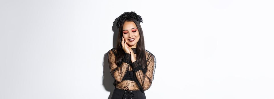 Image of sensual, beautiful asian woman in gothic lace dress and black wreath smiling coquettish, touching face and looking down, standing over white background.