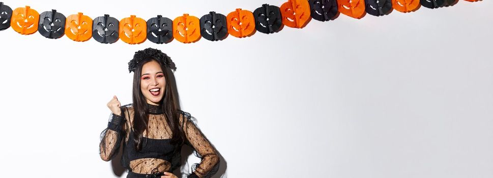 Image of happy asian woman in witch costume having fun on halloween party, saying yes cheerful, standing against pumpkin banners.