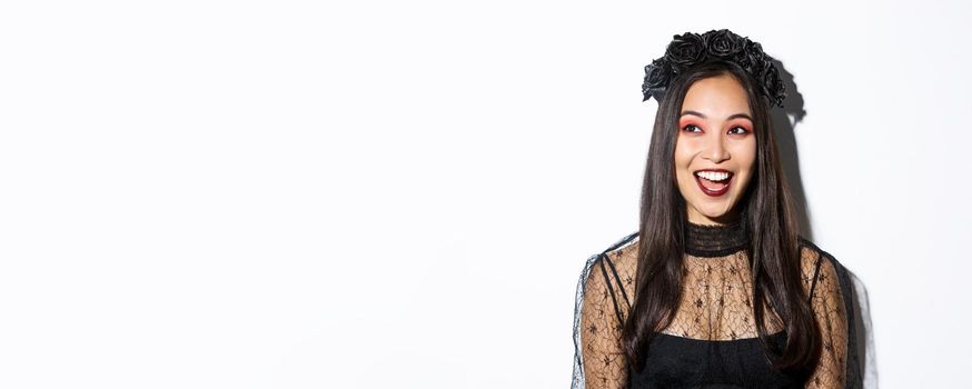 Close-up of happy pretty asian woman in halloween costume looking excited at upper left corner, standing over white background.