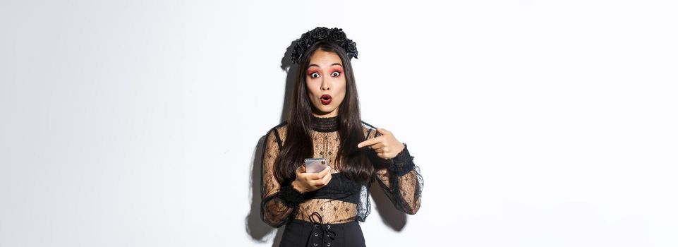 Portrait of amazed asian girl pointing finger at smartphone, showing something about halloween in internet, standing over white background in gothic lace dress.