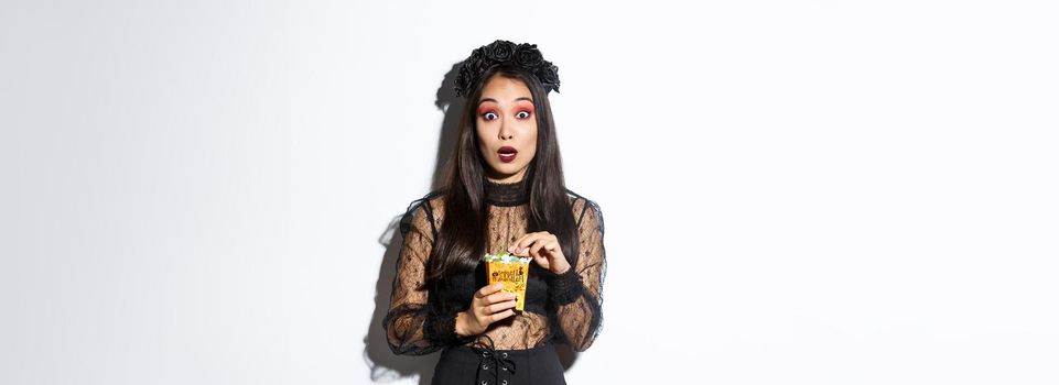 Image of amazed asian woman in witch halloween costume looking at camera while holding sweets in trick or treat bag, standing over white background.