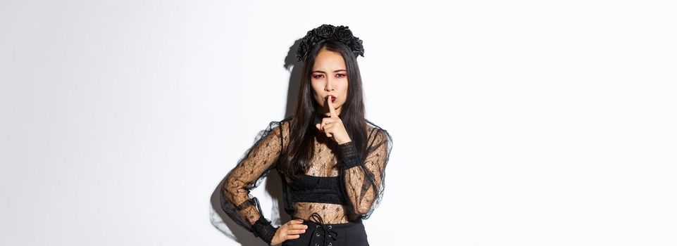 Mysterious asian witch shushing at you with concerned face, hiding secret, tell be quiet. Woman in undead widow costume prepare surprise for halloween, standing over white background.