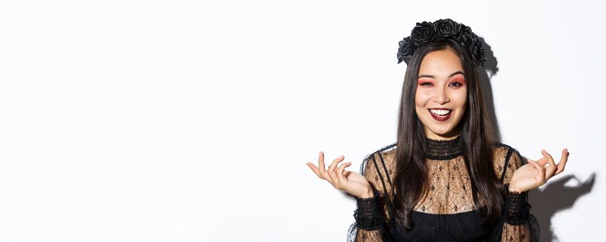 Close-up of sassy pretty asian woman in wicked witch costume making evil laugh and spread hands sideways, standing over white background.