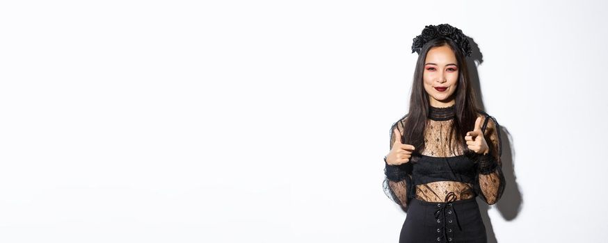 Cheeky and happy attractive woman in witch outfit pointing fingers at camera and smiling, making compliment or praising something good, standing in halloween costume over white background.