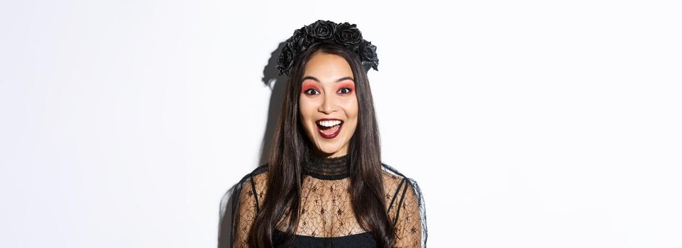 Close-up of enthusiastic asian woman looking amused, wearing witch costume on halloween, standing over white background.