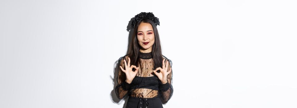 Image of smiling asian cute witch wearing black gothic dress and wreath showing okay gestures in approval, praise something, standing in halloween costume against white background.
