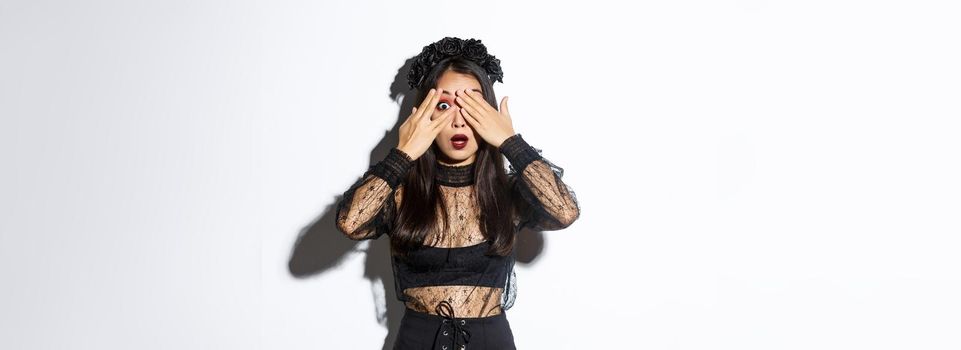 Image of attractive asian girl in halloween costume peeking at something curious, close eyes with fingers but looking through fingers, standing over white background.