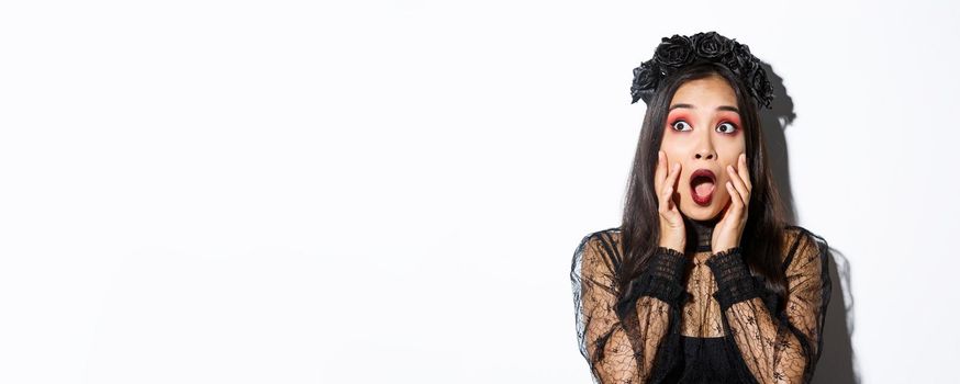 Close-up of scared asian woman in halloween costume of witch looking frightened at upper left corner, gasping and standing ambushed over white background.