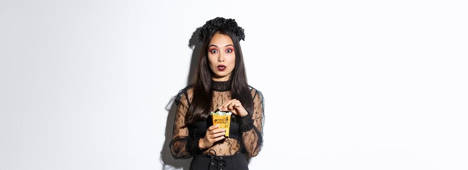 Image of surprised and fascinated asian woman in witch costume, holding sweets gathered during trick or treating, standing over white background.