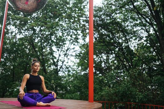 Young woman is practising yoga sitting in the lotus position on a wooden podium in the garden