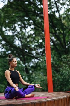 Young woman is practising yoga sitting in the lotus position on a wooden podium in the garden