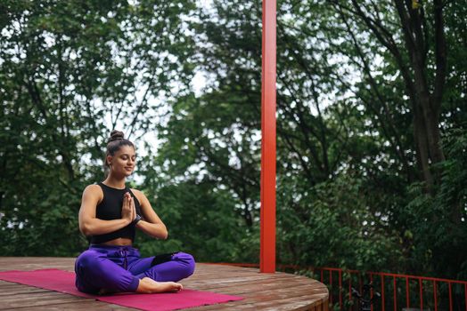 Young woman is practising yoga sitting in the lotus position on a wooden podium in the garden