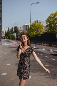 Beautiful woman is walking down the street with smartphone in her hands. Cute girl talking on mobile phone on modern city background