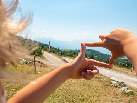 Blond hair boy is framing by fingers wooden tourist sign at gravel road to hill peak. Enjoy trek and funny moment.  Dolomite Alps, Trento region, Italy 