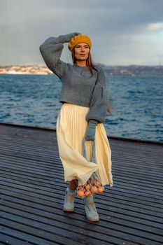 Outdoors fashion portrait of a beautiful middle aged woman walking on the beach. Marine background. Dressed in a stylish warm blue sweater, yellow skirt and beret