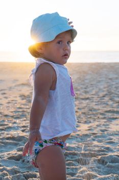 Little blonde girl in a panama hat stands with her back on the beach and looks at the ocean