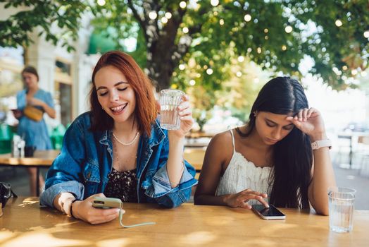 Two female friends browsing their mobile phones while sitting together at city street cafe. Girlfriends ignoring each other while staring in gadgets.
