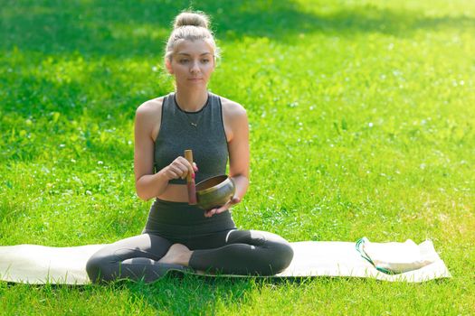 A slender woman in a gray top and leggings, sitting on the green grass in summer, holds a meditation bowl. Copy space