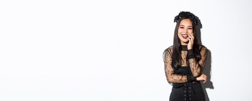 Image of happy good-looking asian woman in gothic dress and black wreath, laughing at halloween party, standing over white background.