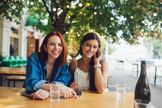 Two female friends browsing in mobile phones while sitting together at city street cafe.