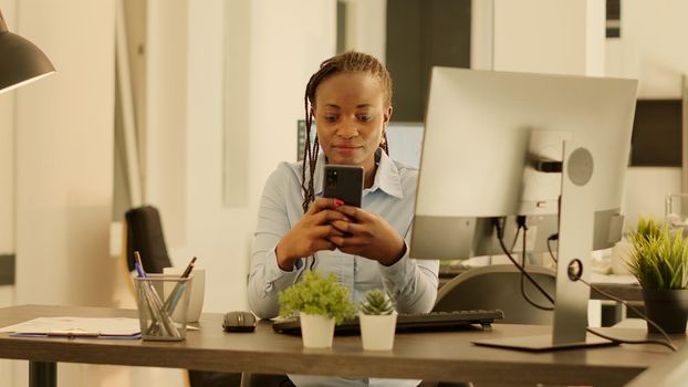 Portrait of startup employee using smartphone at office job during sunset, working on business data report with social media web app. Creating research presentation with mobile phone. Handheld shot.