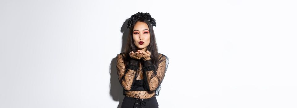 Image of sexy asian woman sending air kiss at camera, ready for halloween party, wearing gothic dress and wreath, celebrating all saints day, standing over white background.