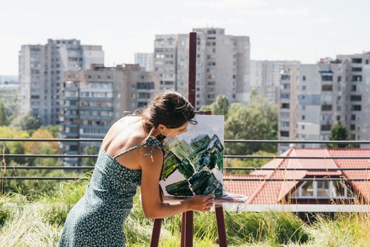 A young beautiful artist paints a picture against the background of the city