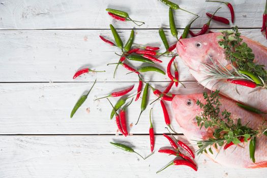 raw fresh pink tilapia fish lies on a board sprinkled with small red and green hot peppers, pink Himalayan salt and garnished with fresh curry sprigs, on white wooden planks. High quality photo